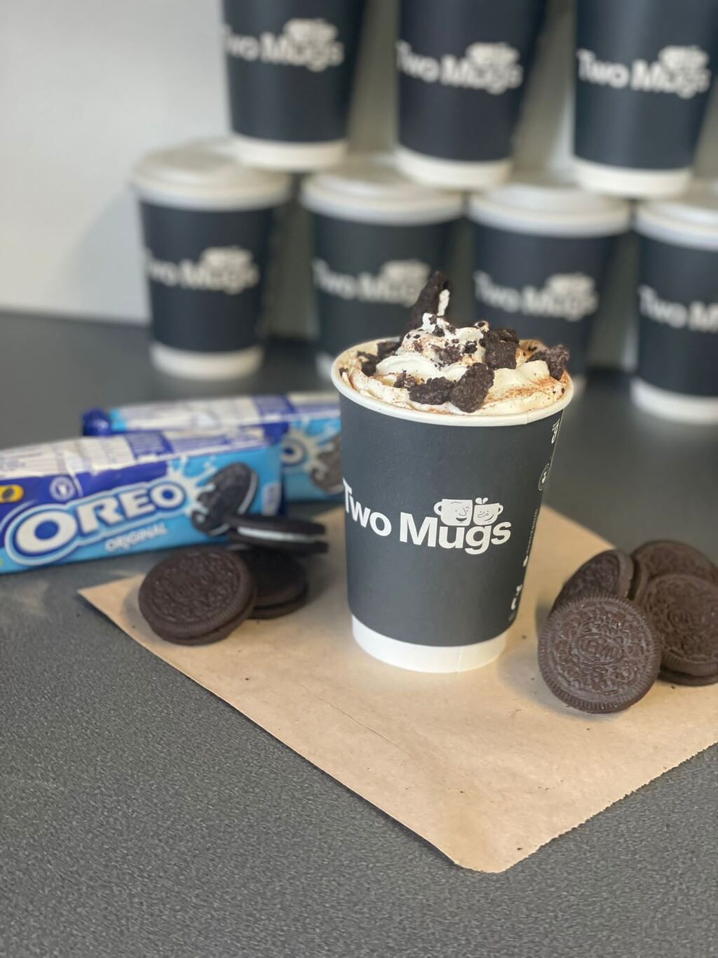 A Two Mugs charcoal cup of hot chocolate topped with cream and crushed Oreo, with biscuits on brown paper and a stack of cups behind