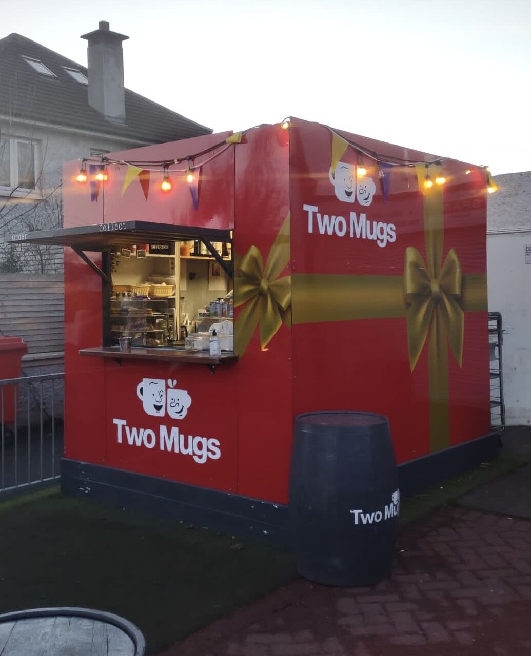 The red Two Mugs container kiosk with its serving hatch, string lights and a barrel in front